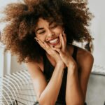 girl with big and long brown natural hair in an afro, sitting in a metal chair, smiling with her eyes closed and her hands on her face with black nail polish and a black tank top shirt