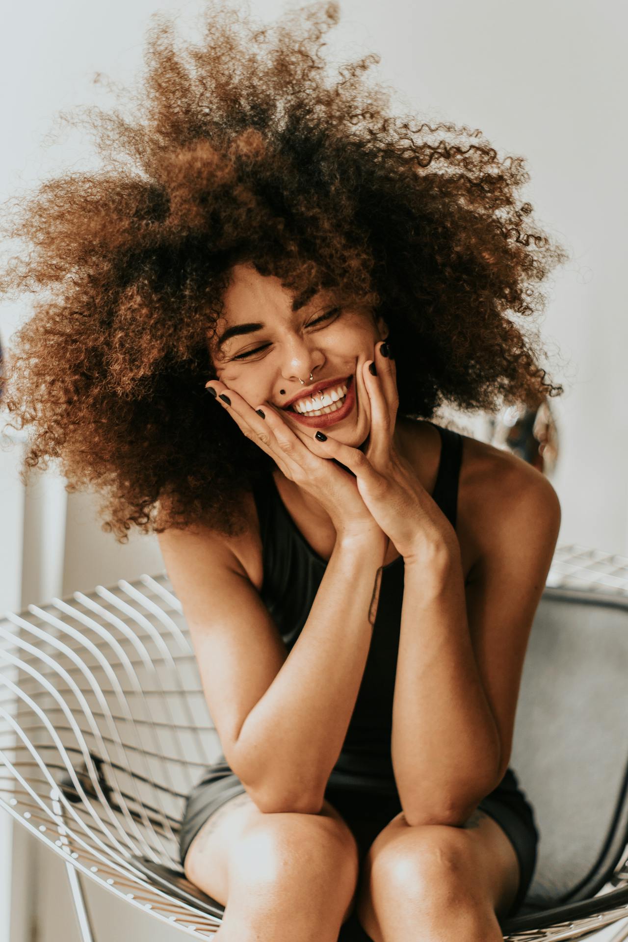 girl with big and long brown natural hair in an afro, sitting in a metal chair, smiling with her eyes closed and her hands on her face with black nail polish and a black tank top shirt