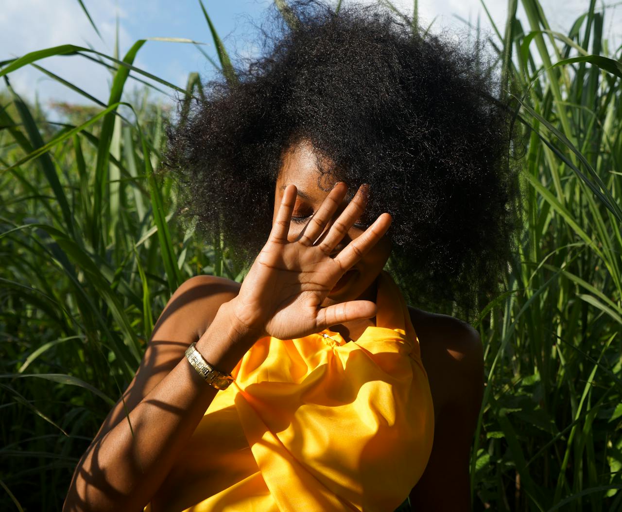 black girl with messy natural black hair in an afro, her hair texture looks like she has heat damage. she has her hand in front of her face, with a bright yellow blouse one and a silver watch