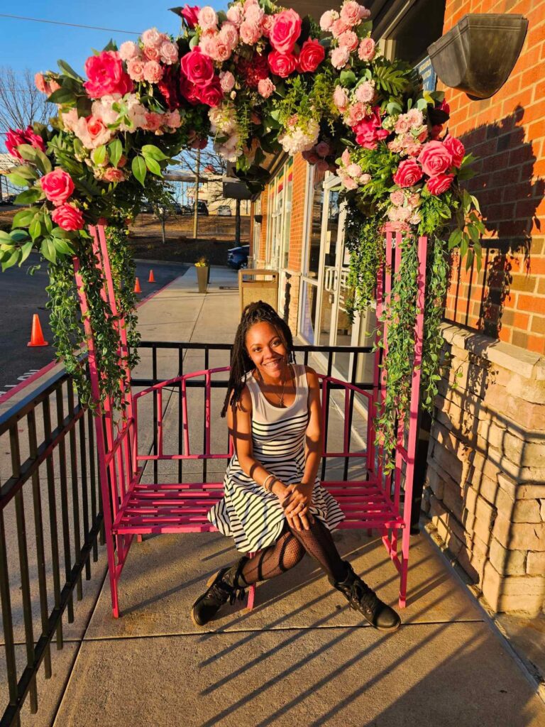 ash randall xo sitting on a pink bench with pink flowers on an arch, dressed in a white and black dress with black fishnets and a black high heel boots